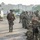 PANAMAX-26: V1/6, Partner forces conduct simulated air raid A group of military personnel in camouflage uniforms are assembled on a tarmac or helipad area near a UH-1 'Huey' utility helicopter. Multiple soldiers wearing tactical gear, helmets, and carrying weapons are visible, with some bearing 'MARINA' (Navy) markings on their vests, suggesting Mexican Naval Infantry involvement. The scene appears to be a pre-mission briefing or staging operation in an urban tropical environment.