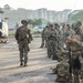 A group of military personnel in camouflage uniforms are assembled on a tarmac or helipad area near a UH-1 'Huey' utility helicopter. Multiple soldiers wearing tactical gear, helmets, and carrying weapons are visible, with some bearing 'MARINA' (Navy) markings on their vests, suggesting Mexican Naval Infantry involvement. The scene appears to be a pre-mission briefing or staging operation in an urban tropical environment.