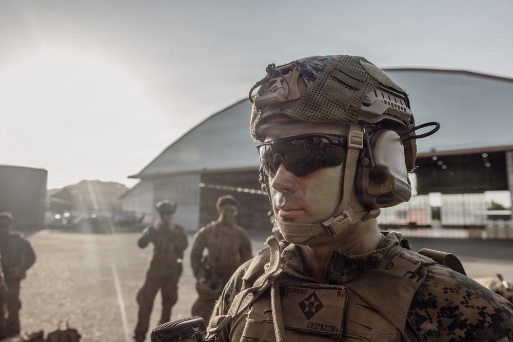 A U.S. Marine in full combat gear is prominently featured in the foreground, wearing a MICH/FAST helmet with communications headset, tactical eyewear, and MARPAT digital camouflage uniform. A name tape reading 'ROBBINS' and a unit patch are visible on the uniform. Additional Marines in similar gear are visible in the background near what appears to be a military airfield hangar.