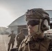 A U.S. Marine in full combat gear is prominently featured in the foreground, wearing a MICH/FAST helmet with communications headset, tactical eyewear, and MARPAT digital camouflage uniform. A name tape reading 'ROBBINS' and a unit patch are visible on the uniform. Additional Marines in similar gear are visible in the background near what appears to be a military airfield hangar.