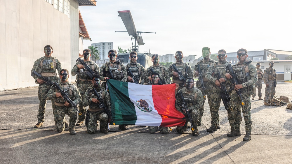 A group of approximately 15 armed military personnel in camouflage uniforms pose for a group photo at what appears to be a military airfield or base. Several members are holding a Mexican flag (green, white, and red tricolor with the national coat of arms), and their uniforms display 'MARINA' patches, identifying them as members of the Mexican Navy (Armada de México). The personnel have tactical face paint applied and are equipped with assault rifles and tactical gear.