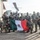 PANAMAX-26: V1/6, Partner forces conduct simulated air raid A group of approximately 15 armed military personnel in camouflage uniforms pose for a group photo at what appears to be a military airfield or base. Several members are holding a Mexican flag (green, white, and red tricolor with the national coat of arms), and their uniforms display 'MARINA' patches, identifying them as members of the Mexican Navy (Armada de México). The personnel have tactical face paint applied and are equipped with assault rifles and tactical gear.