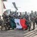 A group of approximately 15 armed military personnel in camouflage uniforms pose for a group photo at what appears to be a military airfield or base. Several members are holding a Mexican flag (green, white, and red tricolor with the national coat of arms), and their uniforms display 'MARINA' patches, identifying them as members of the Mexican Navy (Armada de México). The personnel have tactical face paint applied and are equipped with assault rifles and tactical gear.