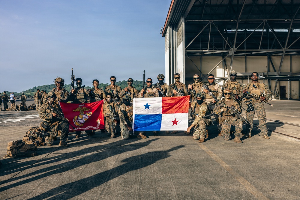 A group of armed military personnel pose for a group photograph on an airfield tarmac in front of a large aircraft hangar. Two flags are prominently displayed: a red U.S. Marine Corps flag on the left and the flag of Panama (white with blue and red stars) in the center. This appears to be a joint military exercise or operation between U.S. Marines and Panamanian security forces.