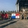 PANAMAX-26: V1/6, Partner forces conduct simulated air raid A group of armed military personnel pose for a group photograph on an airfield tarmac in front of a large aircraft hangar. Two flags are prominently displayed: a red U.S. Marine Corps flag on the left and the flag of Panama (white with blue and red stars) in the center. This appears to be a joint military exercise or operation between U.S. Marines and Panamanian security forces.
