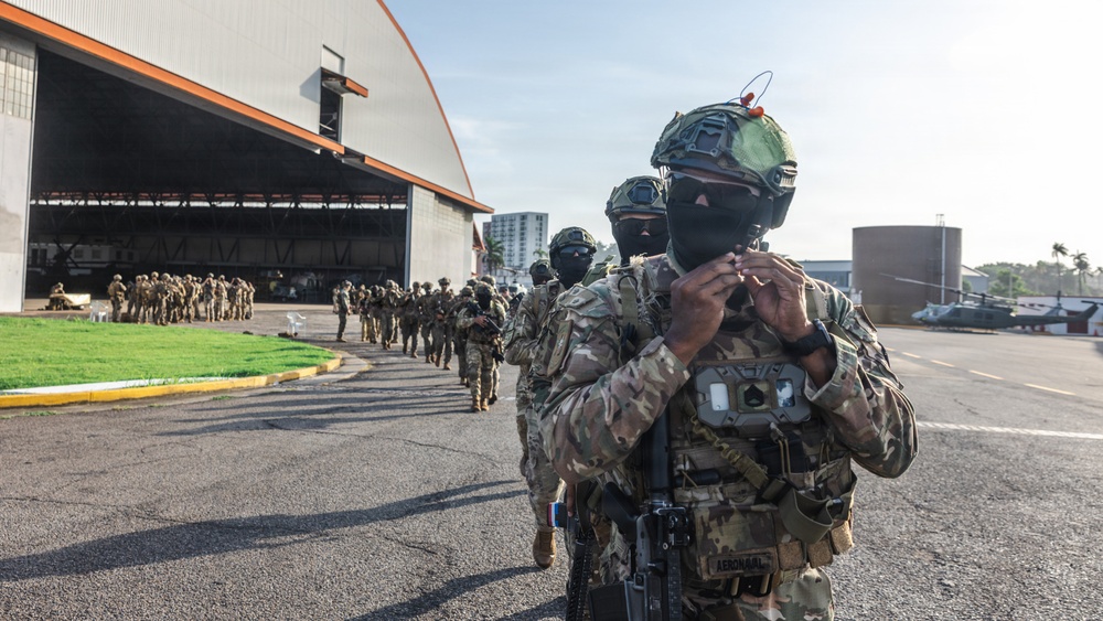 A column of heavily armed and equipped military or special operations personnel marches across an airfield tarmac toward a large aircraft hangar. The lead soldier in the foreground wears full tactical gear including a ballistic helmet with night-vision mount, multicam camouflage, face covering, and a chest-mounted device. A patch or label reading 'AERONAVAL' is visible on the lead soldier's uniform, suggesting a naval aviation or coast guard unit.