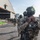 PANAMAX-26: V1/6, Partner forces conduct simulated air raid A column of heavily armed and equipped military or special operations personnel marches across an airfield tarmac toward a large aircraft hangar. The lead soldier in the foreground wears full tactical gear including a ballistic helmet with night-vision mount, multicam camouflage, face covering, and a chest-mounted device. A patch or label reading 'AERONAVAL' is visible on the lead soldier's uniform, suggesting a naval aviation or coast guard unit.