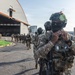 A column of heavily armed and equipped military or special operations personnel marches across an airfield tarmac toward a large aircraft hangar. The lead soldier in the foreground wears full tactical gear including a ballistic helmet with night-vision mount, multicam camouflage, face covering, and a chest-mounted device. A patch or label reading 'AERONAVAL' is visible on the lead soldier's uniform, suggesting a naval aviation or coast guard unit.