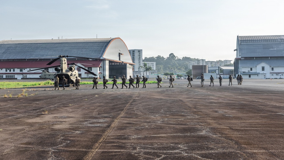 A line of military personnel in full combat gear is boarding or disembarking a CH-47 Chinook helicopter on a tarmac at what appears to be a military airfield. Large aircraft hangars are visible in the background along with urban buildings, suggesting a location near a city. The scene depicts a tactical troop movement or training exercise.