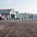 A line of military personnel in full combat gear is boarding or disembarking a CH-47 Chinook helicopter on a tarmac at what appears to be a military airfield. Large aircraft hangars are visible in the background along with urban buildings, suggesting a location near a city. The scene depicts a tactical troop movement or training exercise.