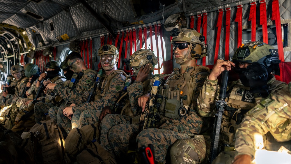 A group of heavily armed military personnel are seated inside what appears to be a CH-47 Chinook helicopter, equipped with full combat gear. The soldiers are wearing a mix of MARPAT (Marine pattern) and MultiCam camouflage uniforms, suggesting a joint or mixed-unit operation. Several personnel have face paint applied, indicating a tactical or special operations mission context.