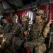 A group of heavily armed military personnel are seated inside what appears to be a CH-47 Chinook helicopter, equipped with full combat gear. The soldiers are wearing a mix of MARPAT (Marine pattern) and MultiCam camouflage uniforms, suggesting a joint or mixed-unit operation. Several personnel have face paint applied, indicating a tactical or special operations mission context.