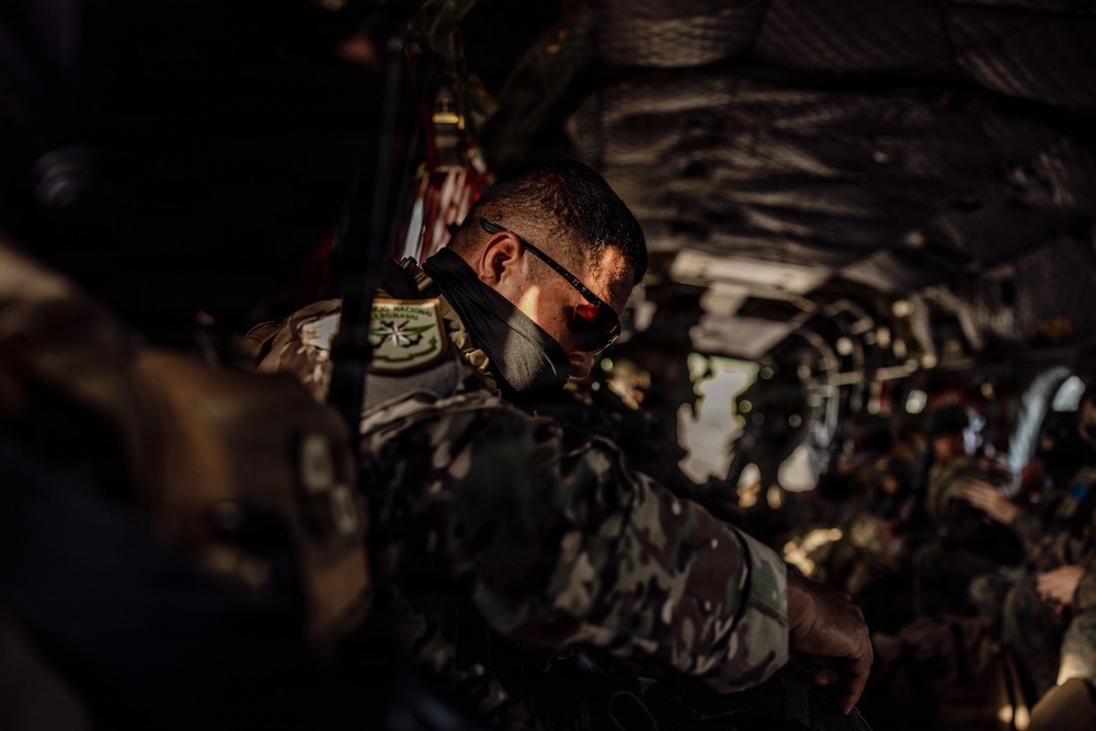 Military personnel are seated inside what appears to be a military transport aircraft or helicopter, packed closely together in full combat gear. The image is taken from inside the aircraft looking toward the rear opening, with dramatic side lighting illuminating the closest soldier. The soldiers are wearing camouflage uniforms and tactical equipment, suggesting an active deployment or training operation.