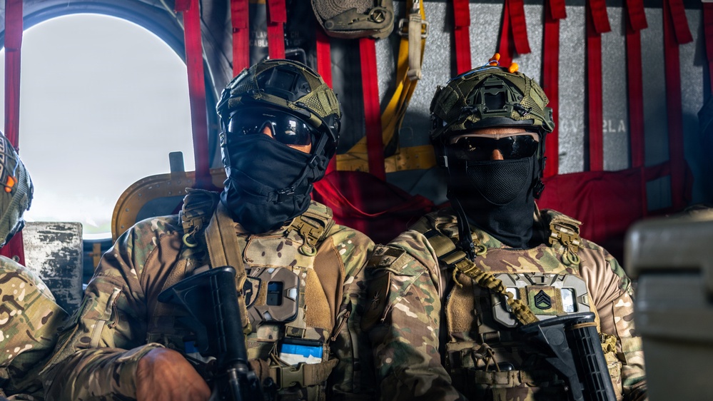 Two heavily equipped military personnel are seated inside what appears to be a military helicopter or transport aircraft. Both individuals are wearing full tactical gear including multicam camouflage uniforms, ballistic helmets, protective goggles, and face coverings. One soldier displays a patch that appears to be the Estonian flag, and the other has visible rank insignia consistent with a sergeant.