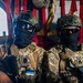 Two heavily equipped military personnel are seated inside what appears to be a military helicopter or transport aircraft. Both individuals are wearing full tactical gear including multicam camouflage uniforms, ballistic helmets, protective goggles, and face coverings. One soldier displays a patch that appears to be the Estonian flag, and the other has visible rank insignia consistent with a sergeant.