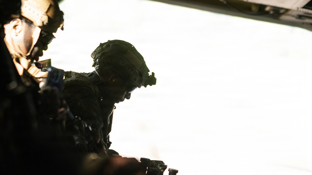 The image shows two heavily equipped military personnel silhouetted against a bright, overexposed background, likely viewed from inside a military aircraft. The figures are wearing tactical helmets with night vision goggle mounts and full combat gear. The strong backlighting creates a dramatic silhouette effect, obscuring most facial and equipment details.