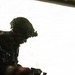 The image shows two heavily equipped military personnel silhouetted against a bright, overexposed background, likely viewed from inside a military aircraft. The figures are wearing tactical helmets with night vision goggle mounts and full combat gear. The strong backlighting creates a dramatic silhouette effect, obscuring most facial and equipment details.