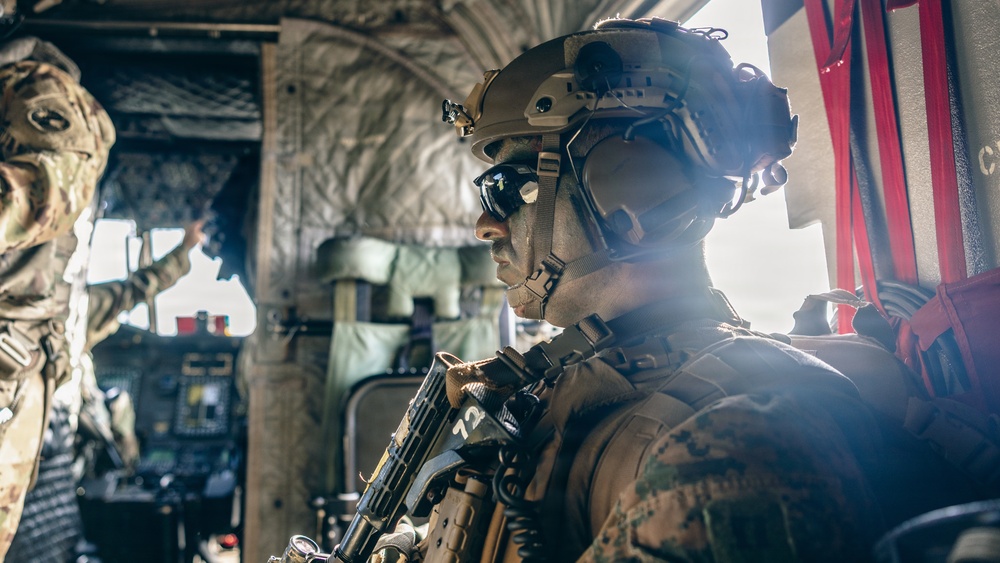 A heavily equipped military operator is seated inside what appears to be a military aircraft or helicopter, wearing full tactical gear including a ballistic helmet and protective eyewear. The soldier is holding a firearm and appears to be in a ready or transit posture. A second soldier is partially visible in the background, gesturing toward the cockpit area.