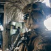 A heavily equipped military operator is seated inside what appears to be a military aircraft or helicopter, wearing full tactical gear including a ballistic helmet and protective eyewear. The soldier is holding a firearm and appears to be in a ready or transit posture. A second soldier is partially visible in the background, gesturing toward the cockpit area.