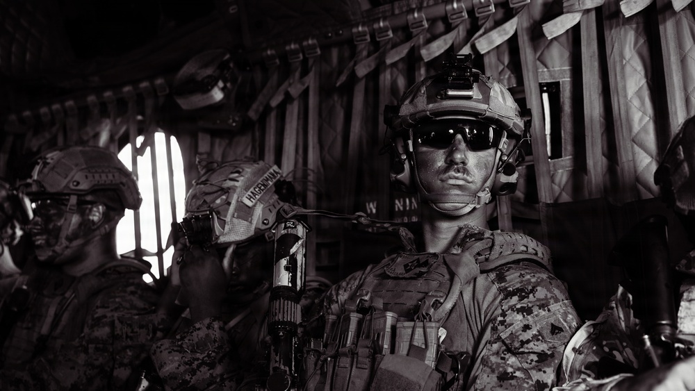 A black-and-white photograph showing military personnel in full combat gear seated inside what appears to be a military transport helicopter. The foreground soldier is equipped with advanced tactical equipment including a helmet-mounted night vision device, tactical sunglasses, and a plate carrier vest. At least two additional soldiers are visible in the background.