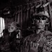 A black-and-white photograph showing military personnel in full combat gear seated inside what appears to be a military transport helicopter. The foreground soldier is equipped with advanced tactical equipment including a helmet-mounted night vision device, tactical sunglasses, and a plate carrier vest. At least two additional soldiers are visible in the background.