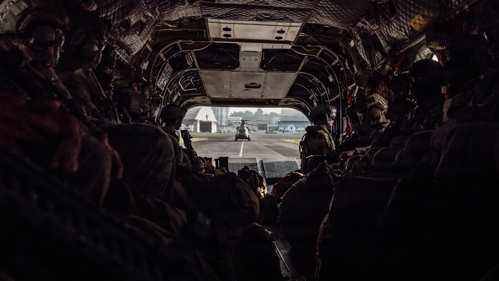 The image is taken from inside a large military helicopter, likely a CH-47 Chinook, looking toward the open rear ramp. Multiple soldiers in full combat gear and helmets are seated along both sides of the aircraft's interior. Through the open ramp, a second helicopter is visible on what appears to be a military airfield tarmac.