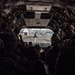 The image is taken from inside a large military helicopter, likely a CH-47 Chinook, looking toward the open rear ramp. Multiple soldiers in full combat gear and helmets are seated along both sides of the aircraft's interior. Through the open ramp, a second helicopter is visible on what appears to be a military airfield tarmac.