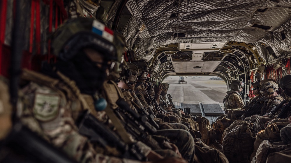 The image shows a group of heavily armed military personnel seated inside what appears to be a CH-47 Chinook helicopter, viewed from the rear cargo bay looking toward the open ramp. Soldiers on the left side display a patch that appears to be the Panamanian flag, suggesting a multinational or joint military operation. Through the open rear ramp, a tarmac or airfield is visible, indicating the aircraft is on the ground or preparing for takeoff.