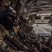 The image shows a group of heavily armed military personnel seated inside what appears to be a CH-47 Chinook helicopter, viewed from the rear cargo bay looking toward the open ramp. Soldiers on the left side display a patch that appears to be the Panamanian flag, suggesting a multinational or joint military operation. Through the open rear ramp, a tarmac or airfield is visible, indicating the aircraft is on the ground or preparing for takeoff.