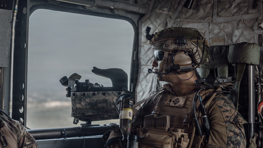 A heavily equipped military service member is seated inside a helicopter, looking out through a large window. The individual is wearing USMC (United States Marine Corps) digital camouflage (MARPAT) uniform with full tactical gear including a combat helmet, communications headset, body armor, and various pouches. A mounted weapon system or optical device is visible in the foreground near the window.