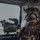 PANAMAX-26: V1/6, Partner forces conduct simulated air raid A heavily equipped military service member is seated inside a helicopter, looking out through a large window. The individual is wearing USMC (United States Marine Corps) digital camouflage (MARPAT) uniform with full tactical gear including a combat helmet, communications headset, body armor, and various pouches. A mounted weapon system or optical device is visible in the foreground near the window.