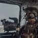A heavily equipped military service member is seated inside a helicopter, looking out through a large window. The individual is wearing USMC (United States Marine Corps) digital camouflage (MARPAT) uniform with full tactical gear including a combat helmet, communications headset, body armor, and various pouches. A mounted weapon system or optical device is visible in the foreground near the window.