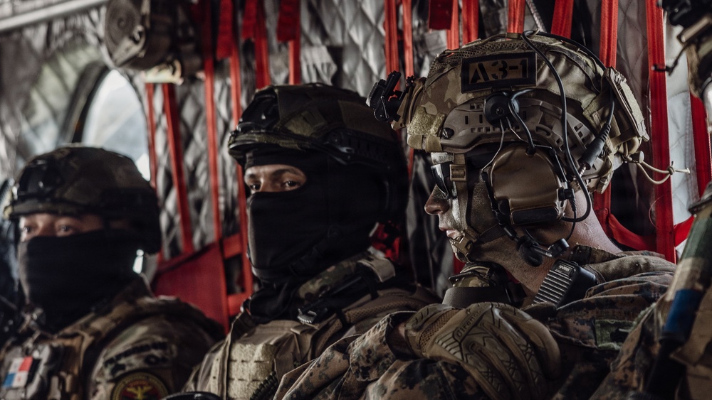 Three military personnel are seated inside what appears to be a military helicopter or transport aircraft, identifiable by the red interior framing and cargo netting. The soldiers are wearing varied tactical gear including helmets, body armor, and communication equipment. At least two different national forces appear to be represented based on visible patches and equipment differences.