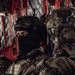 Three military personnel are seated inside what appears to be a military helicopter or transport aircraft, identifiable by the red interior framing and cargo netting. The soldiers are wearing varied tactical gear including helmets, body armor, and communication equipment. At least two different national forces appear to be represented based on visible patches and equipment differences.