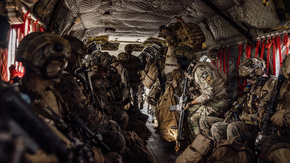The image shows a large group of heavily armed and equipped military personnel seated inside what appears to be a military transport helicopter, likely a CH-47 Chinook, based on the interior configuration. The soldiers are wearing full combat gear including helmets, body armor, tactical vests, and carrying rifles. The aircraft interior features quilted insulation panels, red cargo straps, and a narrow central aisle.