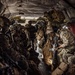 The image shows a large group of heavily armed and equipped military personnel seated inside what appears to be a military transport helicopter, likely a CH-47 Chinook, based on the interior configuration. The soldiers are wearing full combat gear including helmets, body armor, tactical vests, and carrying rifles. The aircraft interior features quilted insulation panels, red cargo straps, and a narrow central aisle.