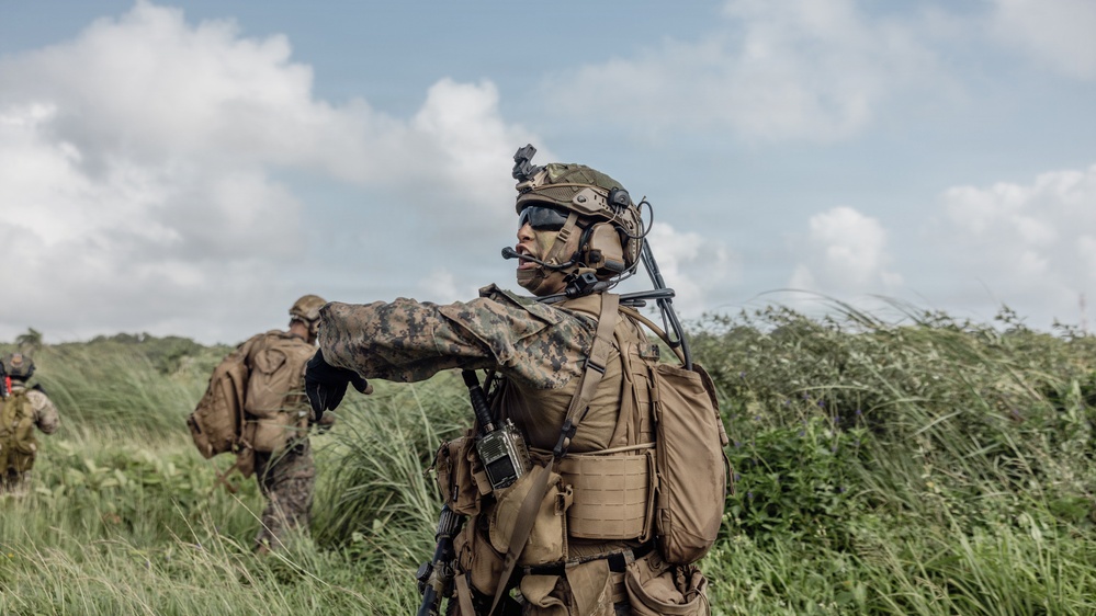 A heavily equipped military service member in the foreground is giving a hand signal while communicating via a tactical headset, with at least two other personnel visible in the background. The setting is an outdoor field environment with dense green vegetation and a partly cloudy sky. The gear and camouflage pattern are consistent with U.S. Marine Corps MARPAT digital camouflage.