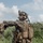 PANAMAX-26: V1/6, Partner forces conduct simulated air raid A heavily equipped military service member in the foreground is giving a hand signal while communicating via a tactical headset, with at least two other personnel visible in the background. The setting is an outdoor field environment with dense green vegetation and a partly cloudy sky. The gear and camouflage pattern are consistent with U.S. Marine Corps MARPAT digital camouflage.