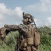 A heavily equipped military service member in the foreground is giving a hand signal while communicating via a tactical headset, with at least two other personnel visible in the background. The setting is an outdoor field environment with dense green vegetation and a partly cloudy sky. The gear and camouflage pattern are consistent with U.S. Marine Corps MARPAT digital camouflage.