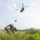 PANAMAX-26: V1/6, Partner forces conduct simulated air raid A military operator in full tactical gear crouches in tall grass while shielding their eyes, observing two CH-47 Chinook tandem-rotor helicopters flying overhead in a low-altitude formation. The setting appears to be a tropical or subtropical environment, indicated by the lush vegetation, palm trees, and a building visible in the background. The scene depicts what appears to be a military exercise or insertion/extraction operation.