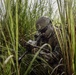 A soldier in full combat gear is positioned low in tall grass, aiming a rifle with an optical scope. The individual is wearing digital camouflage uniform consistent with USMC MARPAT pattern, a tan tactical helmet, and protective eyewear. The scene depicts a concealed tactical position in a dense vegetation environment.