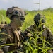 The image shows military personnel in full combat gear positioned in a field of tall green grass, likely during a training exercise or field operation. The foreground soldier wears MARPAT (Marine pattern) camouflage, a tan tactical helmet with communications equipment, tactical vest, and sunglasses with green face paint applied. At least two additional soldiers are visible in the background wearing similar gear.