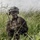 PANAMAX-26: V1/6, Partner forces conduct simulated air raid A fully equipped military service member is positioned low in tall green grass, appearing to communicate via radio handset. The soldier is wearing MARPAT (Marine pattern) camouflage uniform and a tactical helmet with mounted equipment, suggesting U.S. Marine Corps affiliation. The image captures a field training or operational scenario in an outdoor, overcast environment.