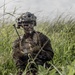 A fully equipped military service member is positioned low in tall green grass, appearing to communicate via radio handset. The soldier is wearing MARPAT (Marine pattern) camouflage uniform and a tactical helmet with mounted equipment, suggesting U.S. Marine Corps affiliation. The image captures a field training or operational scenario in an outdoor, overcast environment.