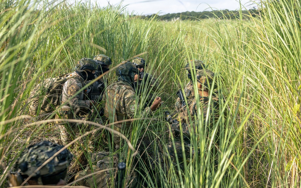 A group of military or special operations personnel in full camouflage gear are concealed within tall grass, appearing to conduct a tactical field operation. The individuals are equipped with helmets, tactical vests, and communications equipment, and appear to be in a briefing or planning huddle. The setting is an open grassy field with dense vegetation providing natural concealment.