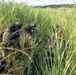 A group of military or special operations personnel in full camouflage gear are concealed within tall grass, appearing to conduct a tactical field operation. The individuals are equipped with helmets, tactical vests, and communications equipment, and appear to be in a briefing or planning huddle. The setting is an open grassy field with dense vegetation providing natural concealment.