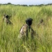 Two armed personnel in military camouflage uniforms and tactical gear are moving through dense, tall green grass in what appears to be a field or savanna environment. Both individuals are wearing helmets and face coverings, with visible unit patches on their sleeves. The image appears to depict a military patrol or training exercise in a tropical or subtropical grassland setting.