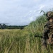 A soldier in full combat gear, wearing MARPAT woodland camouflage, is kneeling in tall grass at the edge of a field while observing a CH-47 Chinook helicopter on or near the ground in the background. Additional personnel are visible near the helicopter in the mid-ground. The scene appears to depict a military helicopter insertion or extraction operation in a lush, green field environment.