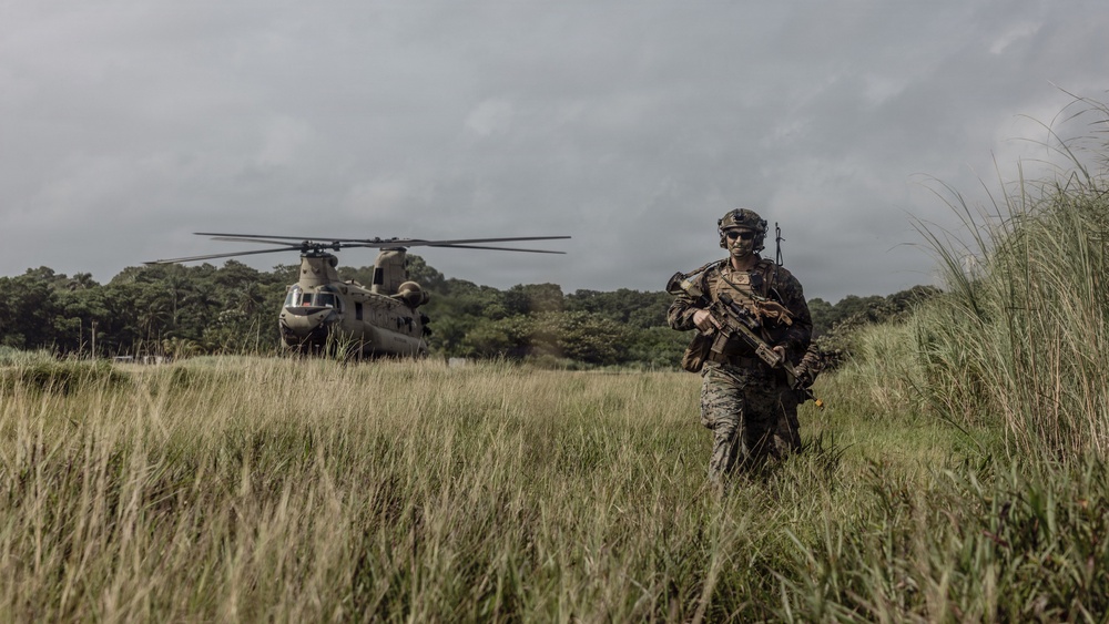 A fully equipped military soldier in camouflage gear walks through a grassy field, moving away from a large tandem-rotor helicopter in the background. The setting appears to be a tropical or subtropical environment with dense tree lines and overcast skies. The scene suggests a recent helicopter insertion or extraction operation.