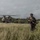 PANAMAX-26: V1/6, Partner forces conduct simulated air raid A fully equipped military soldier in camouflage gear walks through a grassy field, moving away from a large tandem-rotor helicopter in the background. The setting appears to be a tropical or subtropical environment with dense tree lines and overcast skies. The scene suggests a recent helicopter insertion or extraction operation.